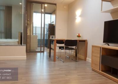 Modern living room with open plan to bedroom, showing a workspace with table and chair, television, and light wooden flooring