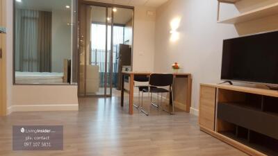 Modern living room with open plan to bedroom, showing a workspace with table and chair, television, and light wooden flooring