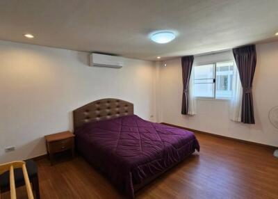 Spacious bedroom with wooden flooring, a double bed, bedside table, chair, air conditioner, ceiling light, window with dark curtains, and a standing fan.