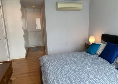 Bedroom with wooden floor, bed with patterned bedding, bedside table with lamp, air conditioning