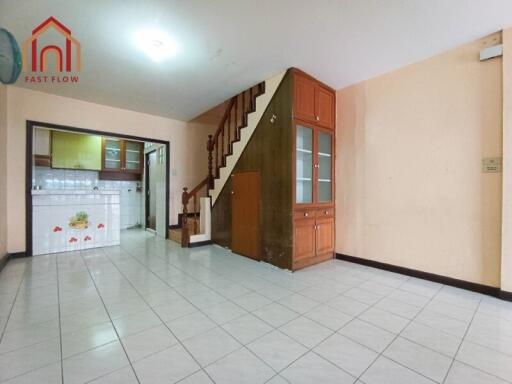 Living room with tiled floor, adjacent kitchen, built-in wooden stair storage, and ample lighting.