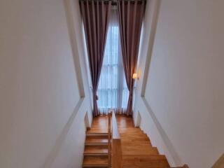 A well-lit staircase with wooden steps, leading to a floor with tall ceiling-height curtains.