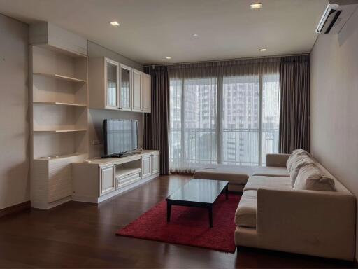 Modern living room with a white couch, built-in shelving, flat-screen TV, and tall windows