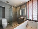 Modern bathroom with glass shower, bathtub, toilet, and large window with blinds