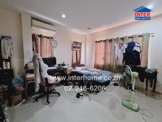 Bedroom with a messy layout including a bed, desk, chair, clothes, and a fan