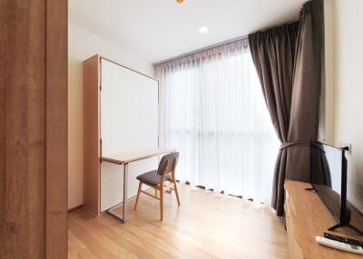 A bright room with wooden flooring, curtains, a foldable desk, and a TV.