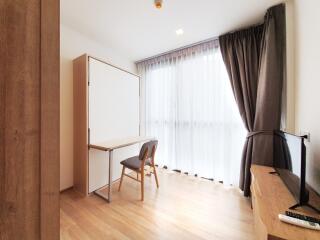 A bright room with wooden flooring, curtains, a foldable desk, and a TV.