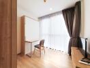 A bright room with wooden flooring, curtains, a foldable desk, and a TV.