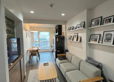 Modern living room with a sofa, coffee table, wall-mounted TV, shelves with framed photos, and dining area in the background.