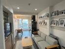 Modern living room with a sofa, coffee table, wall-mounted TV, shelves with framed photos, and dining area in the background.