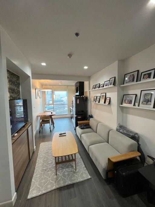 Modern living room with a sofa, coffee table, wall-mounted TV, shelves with framed photos, and dining area in the background.