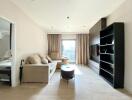 Modern living room with beige sofas, a coffee table, built-in shelves, and a wall-mounted TV, featuring natural light from large windows with beige curtains.