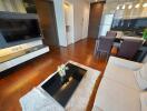 Modern living room with wooden floor, a flat-screen TV, coffee table, and adjoining kitchen