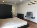 Spacious bedroom with wooden flooring, featuring a large bed, wall-mounted air conditioner, small desk, and modern bathroom in the background.