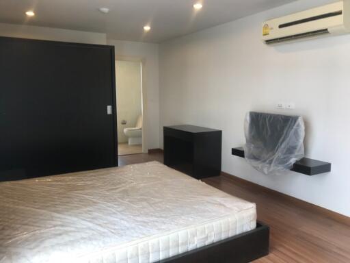 Spacious bedroom with wooden flooring, featuring a large bed, wall-mounted air conditioner, small desk, and modern bathroom in the background.