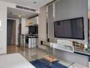 Modern living room with television, glass coffee table, and partial view of kitchen