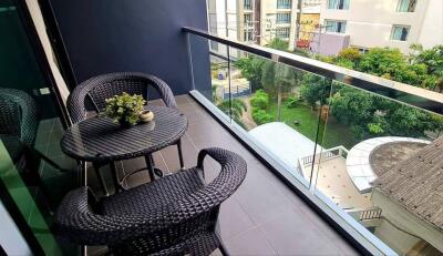 A balcony with two wicker chairs, a table, and a view of a garden and buildings.