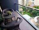 A balcony with two wicker chairs, a table, and a view of a garden and buildings.