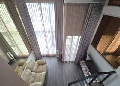 High-ceiling living room with large vertical windows, curtains, beige couch, hardwood floors, and glass staircase