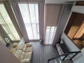 High-ceiling living room with large vertical windows, curtains, beige couch, hardwood floors, and glass staircase