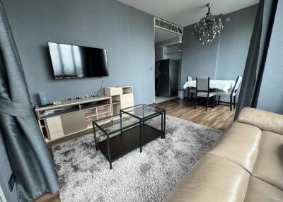 Living room with beige leather sofa, wall-mounted TV, black-glass coffee table, and dining area with chandelier