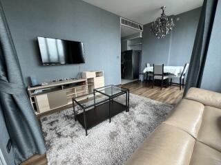 Living room with beige leather sofa, wall-mounted TV, black-glass coffee table, and dining area with chandelier