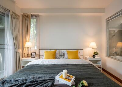 Modern bedroom with a window view, featuring a neatly made bed with yellow pillows, two bedside tables with lamps, and decorative items