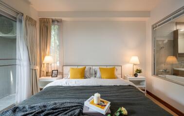 Modern bedroom with a window view, featuring a neatly made bed with yellow pillows, two bedside tables with lamps, and decorative items