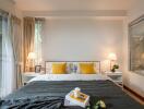 Modern bedroom with a window view, featuring a neatly made bed with yellow pillows, two bedside tables with lamps, and decorative items