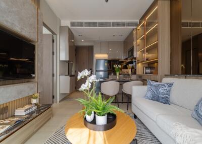 Modern living room with a view into the kitchen, featuring a sofa, coffee table with plants, built-in shelving, and a wall-mounted TV