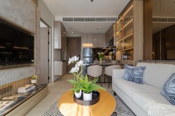 Modern living room with a view into the kitchen, featuring a sofa, coffee table with plants, built-in shelving, and a wall-mounted TV