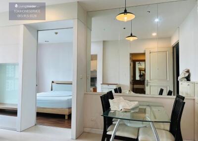 Dining area adjacent to a room with bed, glass table with chairs, and mirror wall.