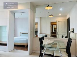 Dining area adjacent to a room with bed, glass table with chairs, and mirror wall.