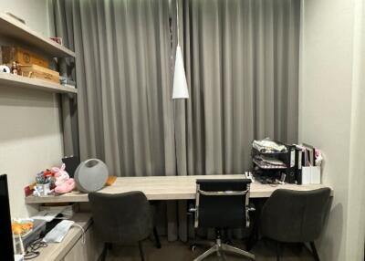 A study room with a large desk and three chairs facing a window with drawn curtains, shelves on the left side, and various items on the desk and shelves