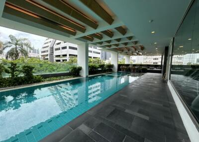 A modern indoor swimming pool area with tiled flooring and ceiling beams, next to large windows overlooking greenery and city buildings.