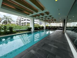 A modern indoor swimming pool area with tiled flooring and ceiling beams, next to large windows overlooking greenery and city buildings.