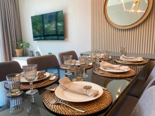 Elegant dining room with a glass table set for a meal, featuring modern décor and a large TV on the wall