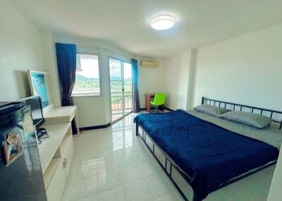 Bright bedroom with a double bed, a TV, a desk with a green chair, and a large window leading to a balcony