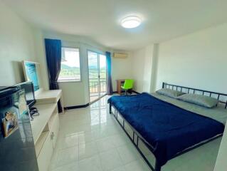 Bright bedroom with a double bed, a TV, a desk with a green chair, and a large window leading to a balcony