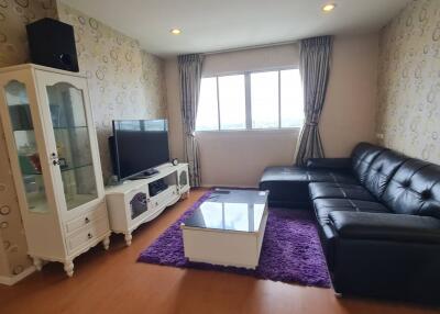 A modern living room with a black leather sectional sofa, glass coffee table on a purple rug, TV with TV stand, and a display cabinet