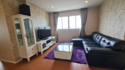 A modern living room with a black leather sectional sofa, glass coffee table on a purple rug, TV with TV stand, and a display cabinet