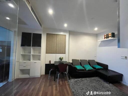 Modern living room with black sofas and teal pillows, wooden flooring, and built-in shelving units