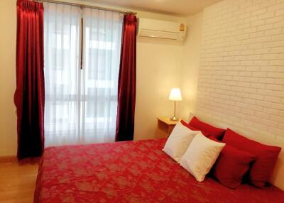 Bedroom with red bedding, white brick wall, bedside table, lamp, and curtains