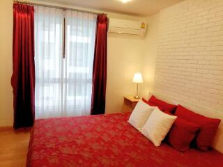 Bedroom with red bedding, white brick wall, bedside table, lamp, and curtains