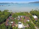 Aerial view of beachfront property with multiple buildings, lush greenery, and a bay with anchored boats