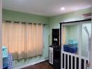 A well-lit bedroom with green walls, a window with beige curtains, and storage furniture