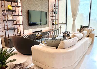 Modern living room with large beige sectional sofa, wall-mounted TV, decorative shelves, and large windows with curtains