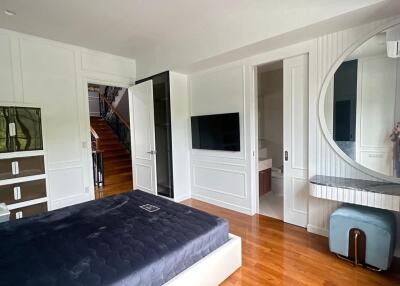 A well-lit modern bedroom with wooden flooring, a large bed, a wall-mounted television, a vanity with a round mirror, and an adjacent en-suite bathroom.