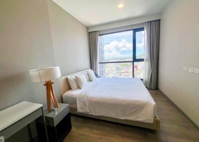 Modern bedroom with city view, featuring a large bed, bedside tables, lamp, and desk.