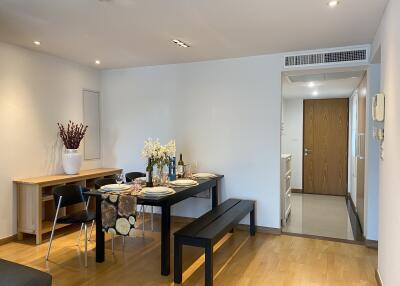 Modern dining area with a black table set for meals, a wooden bench and chairs, decorative flowers, and a sideboard against a light-colored wall.